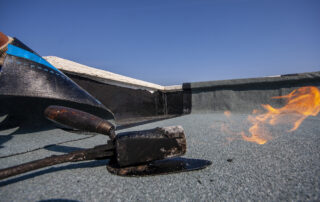 Roofing professional heating and applying a bitumen membrane with a torch, demonstrating the installation context for highperformance asphalt roofing compounds.