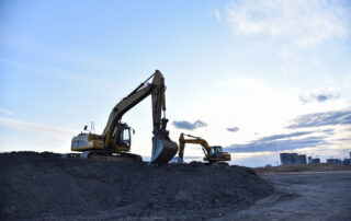 Two excavators at a highway construction site loading old, torn-up road debris at sunset, representing the processing of reclaimed asphalt pavement before applying different types of bitumen additives.