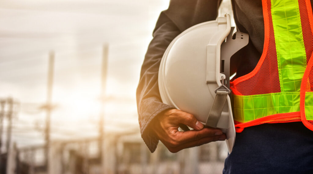 Technician holding white hat safety hard hat sunlight background Construction worker holding a white safety hard hat, representing worker safety and the importance of construction professionals to infrastructure development.