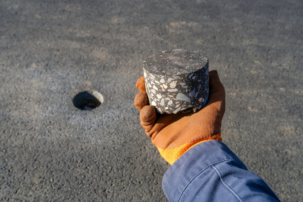 Core sample of road surface material held by a worker in protective glove, with a drilled hole in asphalt. Concept of road repair and material analysis Asphalt core sample illustrating balanced mix design and performance-based asphalt specification testing.